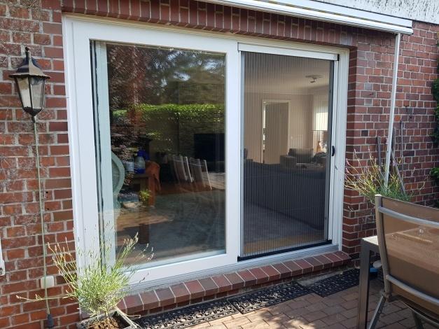 Große weiße Schiebetür mit Insektenschutz und Glasfront in roter Klinkerwand, öffnet den Wohnraum zur Terrasse mit Gartenblick.
