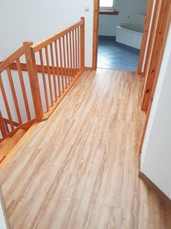 Vinylboden im Obergeschoss Flur mit hellbraunem Vinylboden in Holzoptik, Holzgeländer und Blick ins angrenzende Badezimmer mit blauen Fliesen.