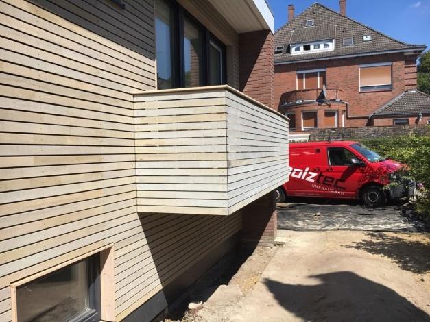 Blick auf die eckige Holzfassade eines modernen Hauses mit großen Fenstern und einem roten Lieferwagen davor. Im Hintergrund ist ein traditionelles Backsteinhaus sichtbar.