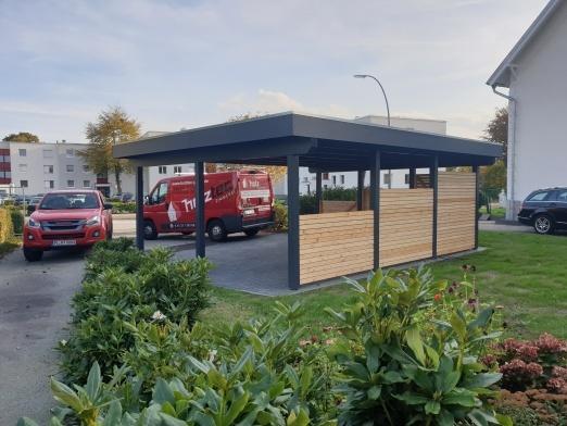 Moderner Carport mit Holzverkleidung in einem Wohngebiet Modern gestalteter Carport mit dunkler Metallstruktur und Holzverkleidung, unter dem zwei Lieferwagen parken. Im Hintergrund sind Wohngebäude und geparkte Autos zu sehen, umgeben von üppigem Grün.